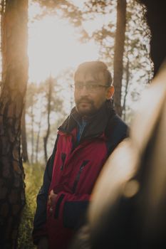 Portrait of a bearded man in a red jacket standing in a sunlit forest during the day.